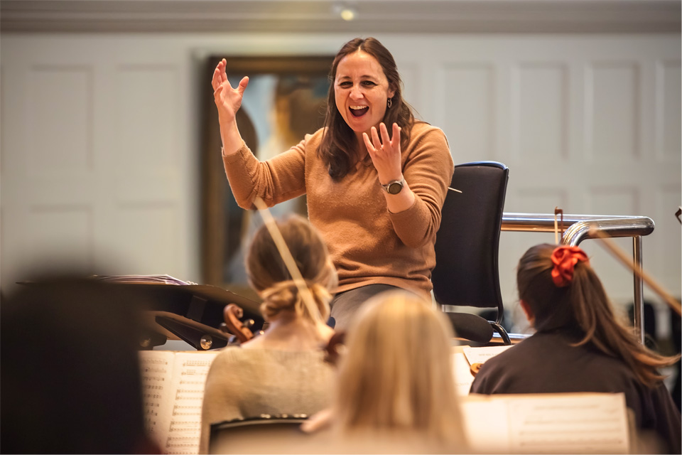 A women, with brown hair, moving her hands, with students sitting in front of her in an orchestra setting, performing on their instruments.